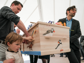 Familie met kleinzoon op de voorgrond draagt een met kindertekeningen versierde kist naar de crematieoven.